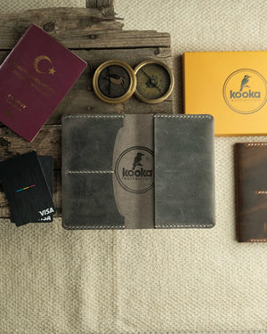 El dikişi gri Crazy Deri pasaport kılıfı, açık düz yatay konumda; ortada belirgin Kooka Leathercraft kabartma logosu, kontrast krem renkli el dikişi kenarlarda net biçimde görünüyor. Deri yüzeyindeki doğal çizikler ve çekme efekti Crazy Deri'nin kendine özgü dokusunu ön plana çıkarıyor.