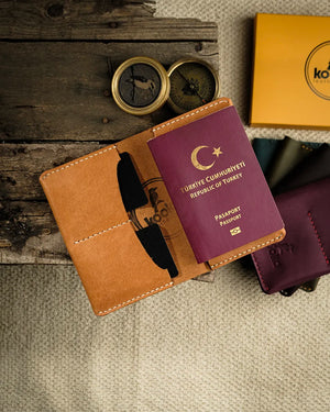 El dikişi deri pasaport kılıfı, camel rengi PUEBLO derisiyle hazırlanmış kooka bifold tasarımında; krem beyazı el dikişi dikişleri, kabartma kookaburra kuş logosu ve düzgün kenar bitişiyle öne çıkan Full Grain (Üst Tabaka) deri yapısı. Açık pozisyonda pasaport yuvası ve kart gözleri net şekilde görülürken arka planda koyu bordo ve koyu yeşil renk seçenekleri de çerçevede yer almaktadır.