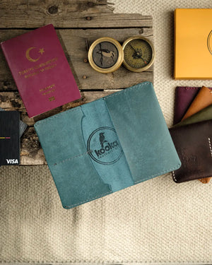 El dikişi deri pasaport kılıfı, mavi Pueblo derisiyle üretilmiş kooka leathercraft imzalı bifold tasarım; yüzeyde doğal mat doku ve belirgin kookaburra kuşu kabartma logosu, temiz dikiş detayları ve özenli kenar işçiliğiyle öne çıkan el yapımı seyahat aksesuar.