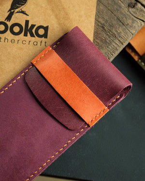 El dikişi deri kalemlik Kooka Leathercraft imzasıyla: mor Pueblo deri gövde üzerinde camel rengi kontrastlı kapama kayışı, düzgün el dikişi ve yumuşak mat yüzey dokusuyla öne çıkan dikey kalemlik detayı.