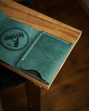 El dikişi deri cüzdan, teal mavi Pueblo derisi ile üretilmiş; bifold tasarımın açık konumda iç kart bölmesi ve el dikişi krem beyazı dikiş detayı ön planda. Pull-up yüzey işleminin yarattığı doğal çizgi izleri ve deri dokusunun zengin teal tonu belirgin biçimde görünürken, kooka leathercraft logolu iç kapak da kareye dahil.