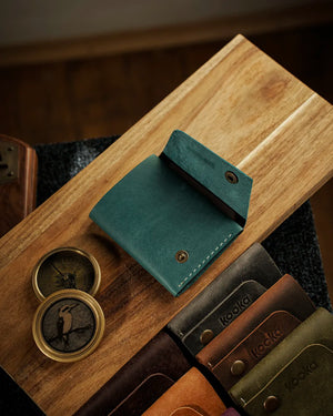 El dikişi deri cüzdan, teal-mavi tonlarında Pueblo derisiyle hazırlanmış kooka STYLE bifold model, antik pirinç çıtçıt detayıyla açık konumda üstten düz çekim; deri yüzeydeki doğal çekme rengi dalgalanmaları ve mat mumsu dokusu belirgin şekilde görünür, seri genelindeki koyu kahverengi, caramel, yeşil ve zeytin rengi modellerin embose kooka logolu yüzeyleriyle birlikte sergilendiği koleksiyon düzenlemesi