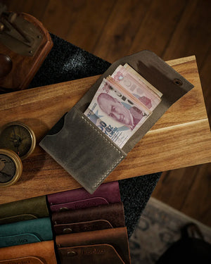 Açık bifold cüzdan tasarımında Crazy Deri yüzeyin gri-yeşil tonları ve doğal çekme renk geçişleriyle Full Grain (Üst Tabaka) derinin karakteri öne çıkan STYLE Dikey Deri Cüzdan, el dikişi bej kontrastlı dikiş sırası ve antik pirinç snap button detayıyla nakit bölmesinde Türk lirası banknotlarını sergilerken altta teal, bordo, kahverengi ve koyu kahve renk seçenekleriyle tüm seri de görünümde yer alıyor; kooka markasının laser kabartma logosu kapak iç yüzeyinde belirgin biçimde konumlanmış.
