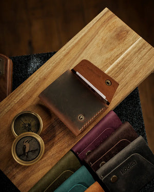 Full Grain (Üst Tabaka) Crazy Deri çoklu renk seçenekleriyle sunulan el dikişi deri cüzdan koleksiyonu; tütün kahvesi açık renkte öne çıkan KOOKA LEATHER STYLE modeli, antik pirinç çıtçıt düğme kapama ve belirgin tahıl dokusuyla fotoğraflanmış, yanında bordo, koyu kahve, teal yeşili, haki ve taba rengi seçenekler sıralanmış — her biri mat, distressed yüzey işçiliği ve lazer kabartma marka logosuyla tamamlanmış