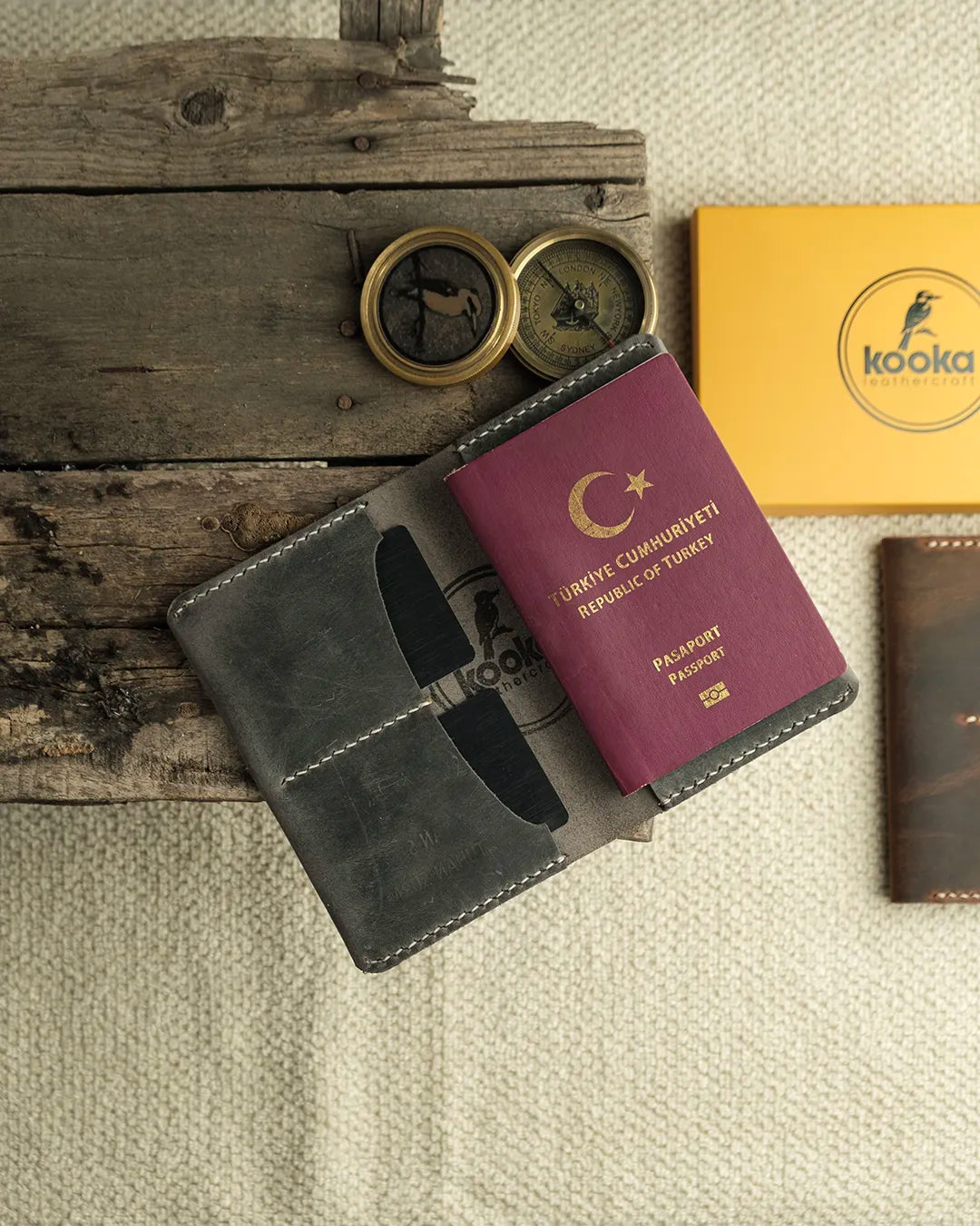 El dikişi gri Crazy Deri pasaport kılıfı, açık konumda pasaport yerleştirilmiş halde üstten düz çekim; yüzeyde doğal çizikler ve pull-up deri karakteristiğiyle zengin doku görünümü, krem kontrast el dikişi tüm kenarlarda belirgin, iki kart gözü ve merkezi pasaport bölmesi net şekilde görünüyor — Kooka Leathercraft imzalı el işçiliği deri aksesuar