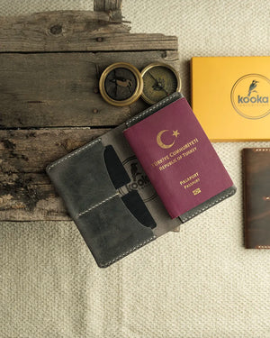 El dikişi gri Crazy Deri pasaport kılıfı, açık konumda pasaport yerleştirilmiş halde üstten düz çekim; yüzeyde doğal çizikler ve pull-up deri karakteristiğiyle zengin doku görünümü, krem kontrast el dikişi tüm kenarlarda belirgin, iki kart gözü ve merkezi pasaport bölmesi net şekilde görünüyor — Kooka Leathercraft imzalı el işçiliği deri aksesuar