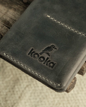 El dikişi gri Crazy Deri pasaport kılıfı üzerinde derin kabartma Kooka marka logosu ve kookaburra kuş figürü detayı — Full Grain (Üst Tabaka) derinin doğal çizik izleri ve mat yüzey dokusuyla birlikte krem renkli el dikişi dikişler tüm kenarlarda belirgin şekilde görünmektedir