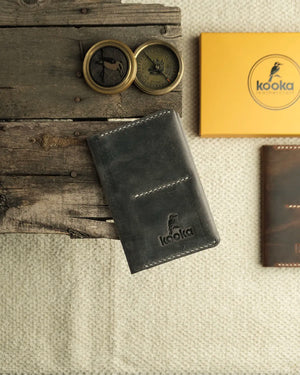 El dikişi pasaport cüzdanı, gri Crazy Deri yüzeyiyle doğal çekme izleri ve doku varyasyonları sergilerken kapağındaki Kooka Leathercraft kabartma logosu zanaatkarlığı ön plana çıkarıyor; beyaz kontrastlı el dikişi kenar işçiliği ve ön kart bölmesiyle sade ama özenli tasarımını kapalı konumda gösteriyor.