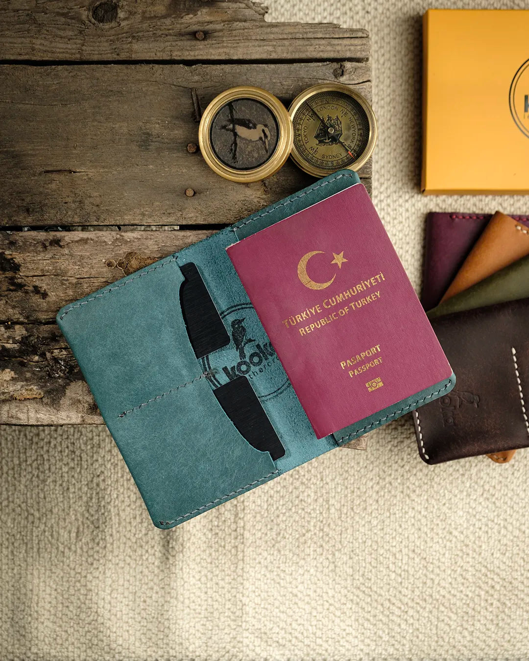 El dikişi deri pasaport kılıfı, mavi Pueblo deri yüzeyiyle açık konumda sergileniyor; sol iç bölmede iki kart yuvası ve sağda pasaport cebi net biçimde görülüyor. Kooka imzalı kookaburra kuşu kabartma logolu bu bifold tasarım, mat ve doğal dokulu Pueblo deri yüzeyi ile elle yapılmış temiz dikiş işçiliğini ön plana çıkarıyor.