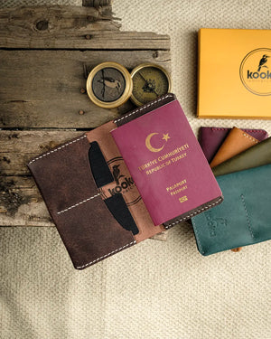 El dikişi deri pasaport kılıfı, Pueblo deri kahverengi rengiyle mat, hafif grenli yüzey dokusu ve belirgin beyaz el dikişi detaylarıyla öne çıkıyor; kooka markasının kabartma logosu iç bölmede yer alıyor. Bifold tasarım, tek pasaport gözü ve iki kart yuvası sunan bu cüzdan, açık konumda ve içinde pasaport görünürken işçilik kalitesini sergiliyor. Arka planda teal ve mürdüm renk seçenekleriyle birlikte sunulan ürün, tam deri el işçiliği seyahat aksesuar koleksiyonunun bir parçasıdır.
