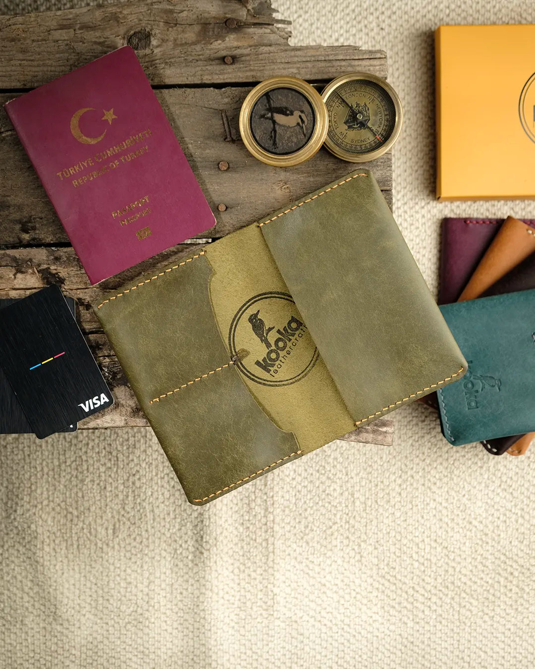 El dikişi deri pasaport kılıfı, zeytin yeşili PUEBLO derisiyle üretilmiş; mat, hafif kadifemsi yüzey dokusu ve altın sarısı el dikişleriyle öne çıkan kooka imzalı bifold tasarım. Kapağın ortasında kabartma kookaburra logosu, temiz kenar bitişleri ve pasaport ile kart gözleriyle tam görünümde sunuluyor.