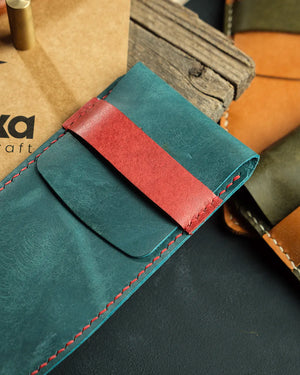 El dikişi deri kalemlik, teal mavi Pueblo deri gövde üzerinde kırmızı kontrastlı kilit askısı ve eşleşen kırmızı el dikişiyle öne çıkan Kooka Leathercraft imzalı dikey kalemlik; deri yüzeyindeki doğal çizikler ve mat-parlak geçişli doku, özgün el işçiliğini yansıtıyor.