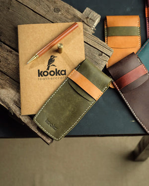 el dikişi deri kalemlik, zeytin yeşili ve camel rengi PUEBLO deri üst tabaka tam tahıl dokusuyla işlenmiş Kooka Leathercraft imzalı dikey kalemlik — krem rengi kontrastlı el dikişi, camel rengi deri kapama askısı ve ön yüzeyde kabartma logo detayıyla birden fazla renk seçeneği bir arada