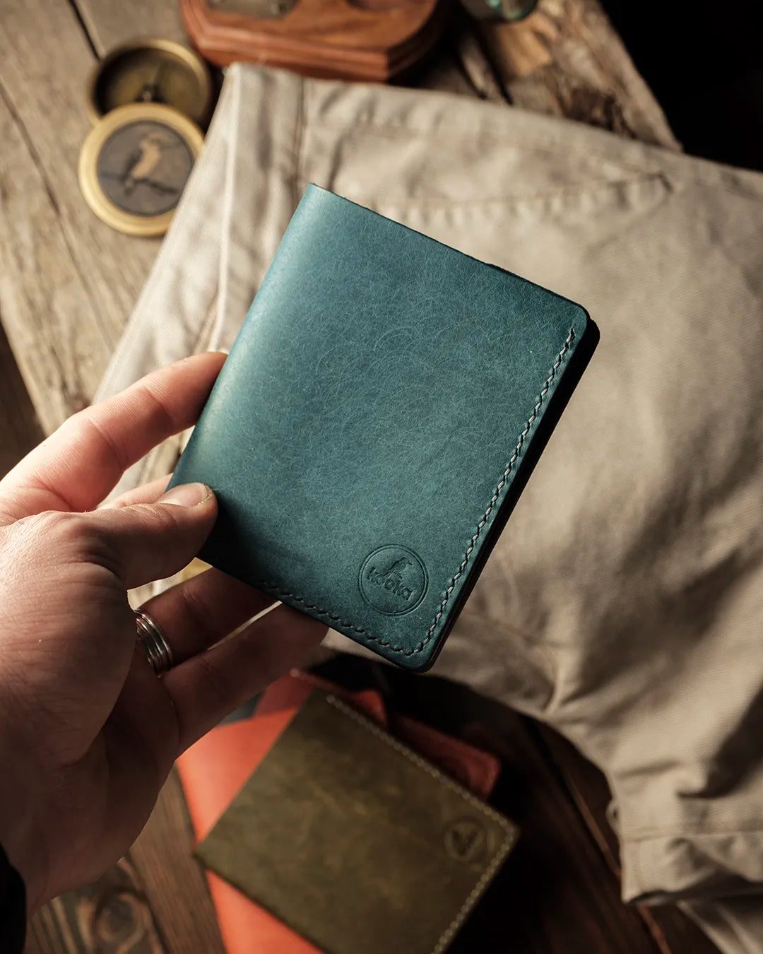El yapımı hakiki deri dikey cüzdan, petrol mavisi PUEBLO deri yüzeyi ve kenar boyunca ton sür ton el dikişi ile kapalı halde elde tutulurken; ön alt köşede kabartma Kooka Leather logosu, avuç içine sığan kompakt THAN modeli