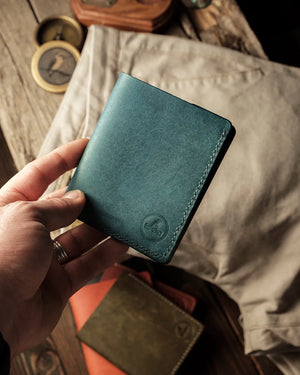 El yapımı hakiki deri dikey cüzdan, petrol mavisi PUEBLO deri yüzeyi ve kenar boyunca ton sür ton el dikişi ile kapalı halde elde tutulurken; ön alt köşede kabartma Kooka Leather logosu, avuç içine sığan kompakt THAN modeli