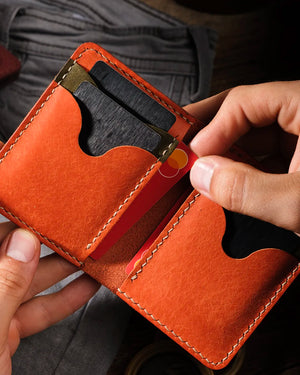 Hands holding open Kooka OZN bifold leather wallet in cognac full-grain vegetable-tanned leather, demonstrating card insertion into scallop-cutout card slot; interior reveals multiple cards including a Mastercard, with cream saddle stitching and warm directional lighting highlighting natural grain texture.