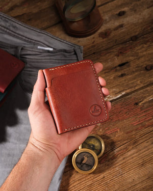 Kooka OZN el yapımı tam dane deri bifold cüzdan, bir erkek eli tarafından tutuluyor; koyu ahşap yüzey üzerinde vintage pirinç pusula ve diğer renk varyantları arka planda görünüyor. Konyak/kestane kahvesi taba rengi, krem ekru el dikişi ve kabartma kingfisher logosu öne çıkıyor.