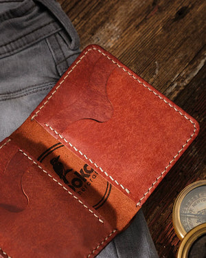 Kooka OZN el yapımı bifold deri cüzdan içi detay — cognac taba rengi tam tahıllı Pueblo deri yüzeyde doğal deri damarları ve pull-up renk varyasyonu belirgin; krem/ekru renkli el dikişi saddle stitch çizgileri ve Kooka marka logosu (kuş motifi) yakın çekimde görünüyor. Arka planda gri denim kumaş ve vintage pirinç pusula ile rustik ahşap zemin, heritage zanaatkarlık estetiğini tamamlıyor.