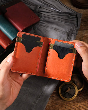 Kooka OZN el yapımı deri bifold cüzdan, iki el tarafından açık tutularak iç kısmı sergileniyor; dalgalı scallop kesimli kart yuvaları, krema rengi el dikişi ve zengin turuncu-cognac tam dane deri iç yüzey görünüyor. Arka planda koyu ahşap zemin üzerinde teal, bordo ve siyah renk varyantları, gri denim pantolon ve pirinç pusula prop olarak konumlandırılmış.