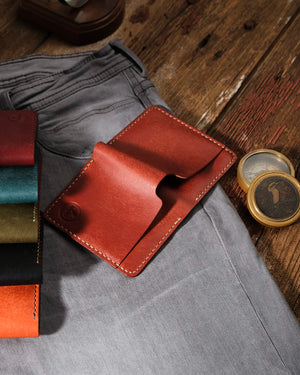 Kooka OZN el yapımı tam dane deri bifold cüzdan, cognac/kestane taba rengi, krem saddle dikişli, vintage pirinç pusula ve gri denim pantolon ile rustik ahşap zemin üzerinde yaşam tarzı düzenlemesi — renk serisi varyantları (bordo, teal, haki, siyah, turuncu) eşliğinde sergileniyor.