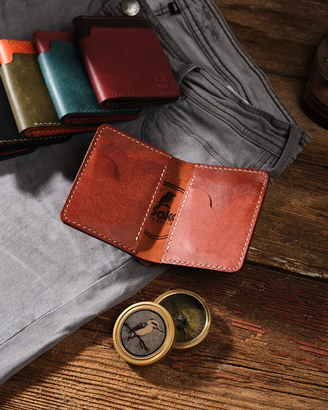 Kooka OZN bifold deri cüzdan açık pozisyonda, cognac/kestane rengi tam tahıl bitkisel sepilenmiş deri, krem el dikişi ve marka logosu ile birlikte rustik ahşap yüzey üzerinde; arka planda zeytin yeşili, teal, bordo ve kahverengi renk varyantları ile gri kot pantolon ve pirinç pusuladan oluşan Heritage-craft düzenlemesi.