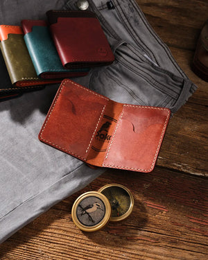 Kooka OZN bifold deri cüzdan açık pozisyonda, cognac/kestane rengi tam tahıl bitkisel sepilenmiş deri, krem el dikişi ve marka logosu ile birlikte rustik ahşap yüzey üzerinde; arka planda zeytin yeşili, teal, bordo ve kahverengi renk varyantları ile gri kot pantolon ve pirinç pusuladan oluşan Heritage-craft düzenlemesi.