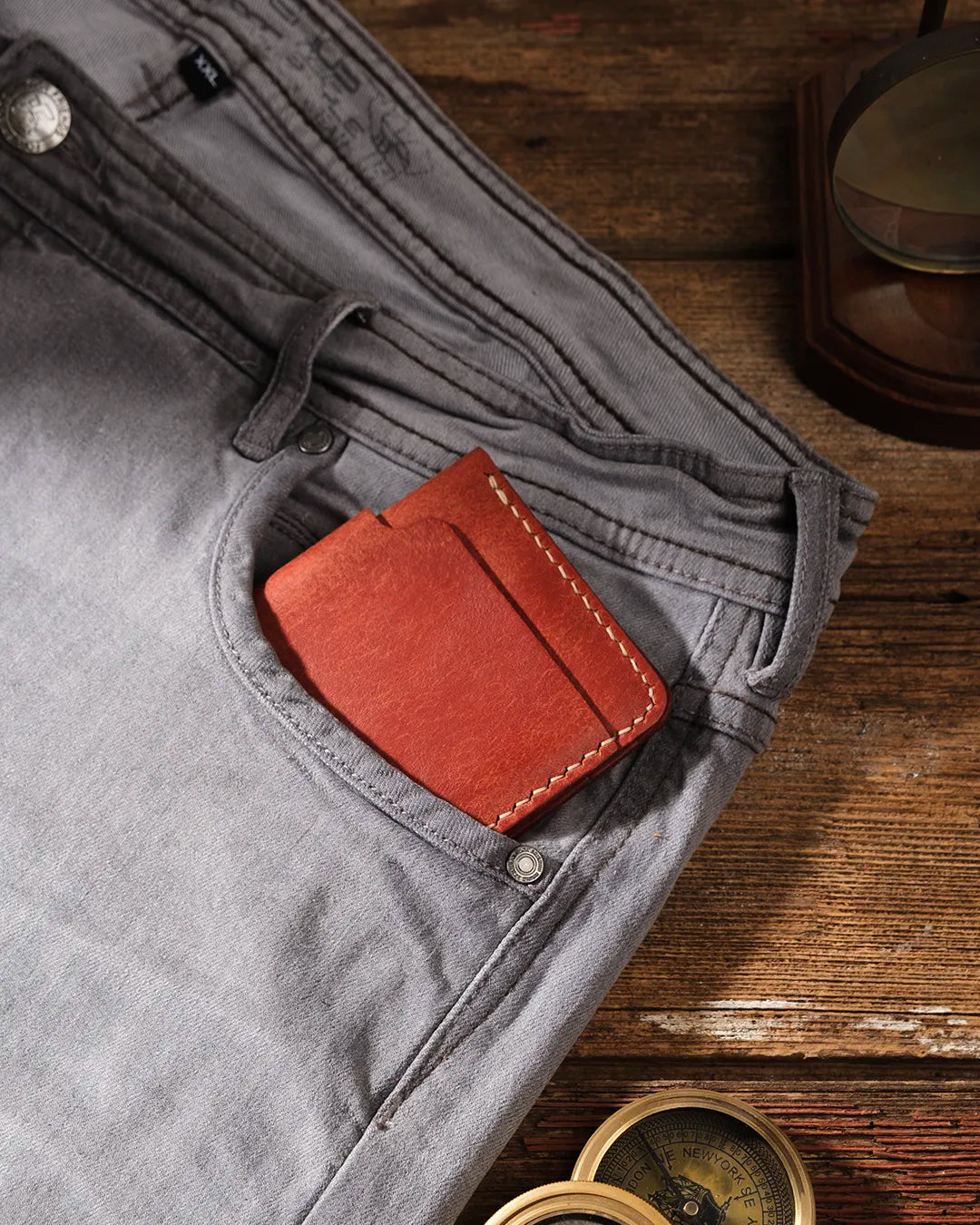 Kooka OZN el yapımı tam dane deri bifold cüzdan, cognac/taba renginde, gri denim pantolonun ön cebinden hafifçe çıkıntı yaparak sergileniyor; arka planda rustik koyu ahşap zemin ve pirinç pusula aksesuarıyla maskülen, maceracı bir atmosfer yaratılmış.