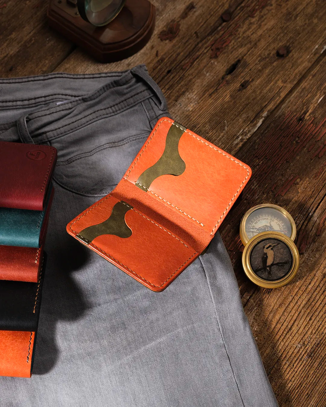 Kooka OZN bifold cüzdan açık pozisyonda, turuncu Pueblo deri iç yüzey ve olive yeşili kart yuvaları görünür şekilde; gri denim pantolon, vintage pirinç pusula ve serinin diğer renk varyantlarıyla birlikte rustik ahşap zemin üzerinde stil düzenlemesi.
