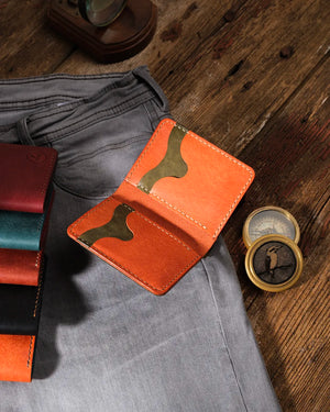 Kooka OZN bifold cüzdan açık pozisyonda, turuncu Pueblo deri iç yüzey ve olive yeşili kart yuvaları görünür şekilde; gri denim pantolon, vintage pirinç pusula ve serinin diğer renk varyantlarıyla birlikte rustik ahşap zemin üzerinde stil düzenlemesi.