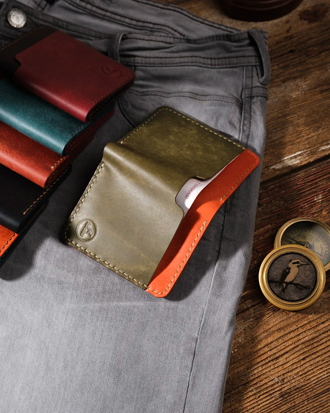 Kooka OZN El Yapımı Deri Bifold Cüzdan – Olive Yeşil & Turuncu İki Tonlu, gri denim pantolonun ön cebinden kart çıkarılırken gösteriliyor; arka planda burgundy, teal ve siyah renk varyantları ile pirinç pusula aksesuarı, rustik ahşap zemin üzerinde sanatsal kompozisyon