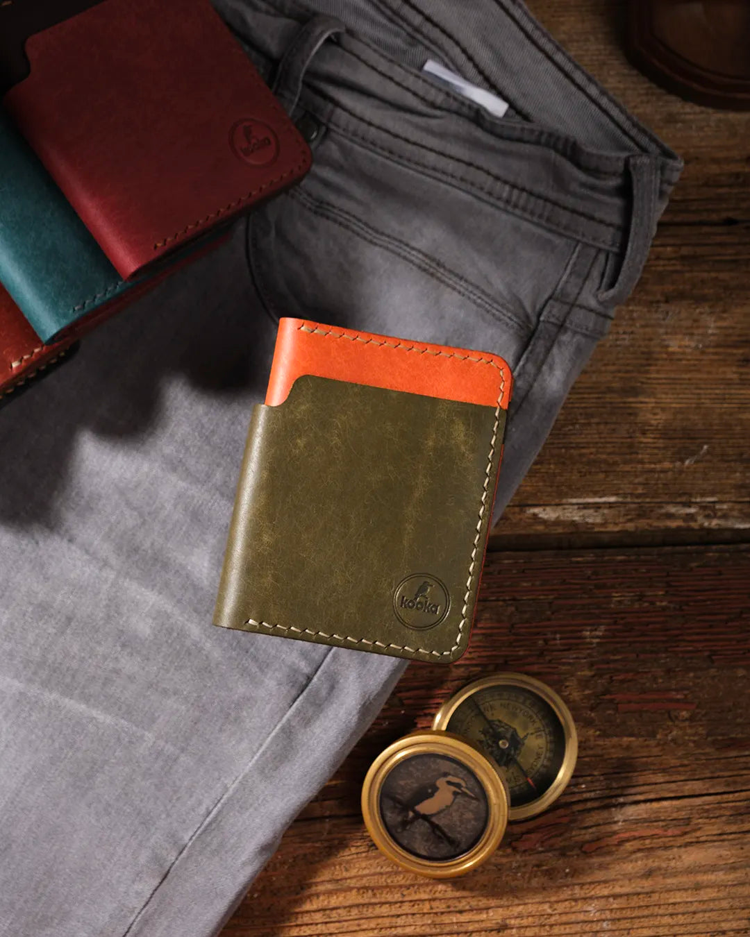 Kooka OZN bifold deri cüzdan, olive yeşili ve turuncu iki tonlu Pueblo deri kombinasyonuyla gri kanvas pantolonun ön cebine yaslanmış şekilde rustik ahşap yüzey üzerinde sergileniyor. Ön yüzde emboss Kooka logosu ve krem renkli el dikişi detayları belirgin. Sahne aksesuarları: teal, bordo ve kahverengi renk varyantları sol üstte gruplu, sağ altta pirinç vintage pusula (kuş figürlü). Maskülen, maceracı ve zanaatkâr atmosfer.