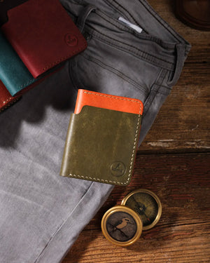 Kooka OZN bifold deri cüzdan, olive yeşili ve turuncu iki tonlu Pueblo deri kombinasyonuyla gri kanvas pantolonun ön cebine yaslanmış şekilde rustik ahşap yüzey üzerinde sergileniyor. Ön yüzde emboss Kooka logosu ve krem renkli el dikişi detayları belirgin. Sahne aksesuarları: teal, bordo ve kahverengi renk varyantları sol üstte gruplu, sağ altta pirinç vintage pusula (kuş figürlü). Maskülen, maceracı ve zanaatkâr atmosfer.