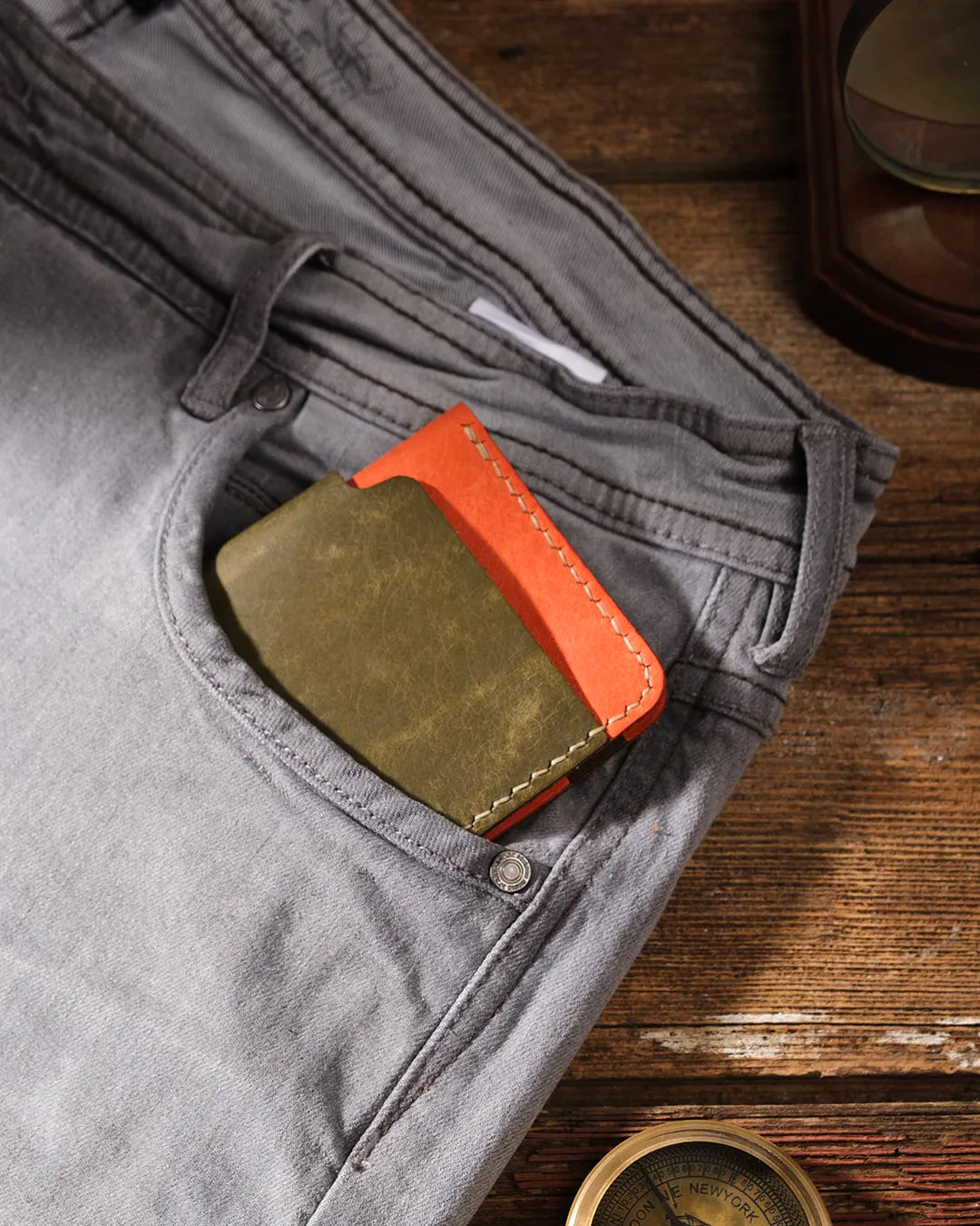 Kooka OZN bifold cüzdan olive yeşil ve turuncu iki tonlu Pueblo derisiyle, gri kanvas pantolonun ön cebinden taşarken görünüyor; arka planda vintage pirinç pusula ve ahşap yüzey ile rustik, maceraperest atmosfer.