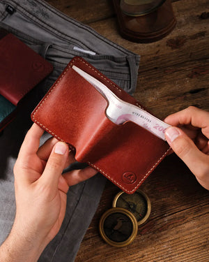 Kooka OZN el yapımı tam dane deri bifold cüzdan, taba/konyak rengi Pueblo derisiyle üretilmiş; iki el tarafından açık tutulurken içinden banknot çıkarılıyor. İçeride krem rengi el dikişi ve scallop kart yuvaları görünür. Arka planda pirinç pusula, kum saati, gri pantolon ve farklı renk varyantları ile rustik ahşap zemin.
