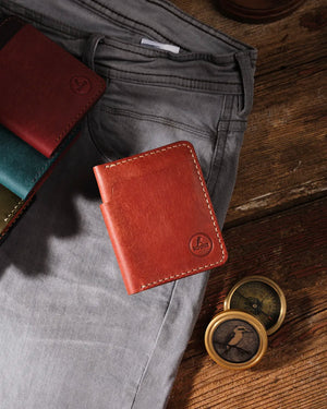Kooka OZN bifold cüzdan, taba/konyak tam dane vegetable-tanned deriden el yapımı; krem rengi el dikiş detayları ve kabartma Kooka logosuyla birlikte gri denim pantolon üzerinde düz yatış kompozisyonuyla sergileniyor. Arka planda burgundy, teal ve olive renk varyantları ile pirinç vintage pusula aksesuarı, rustik koyu ahşap zemin üzerinde heritage-craft atmosfer yaratıyor.