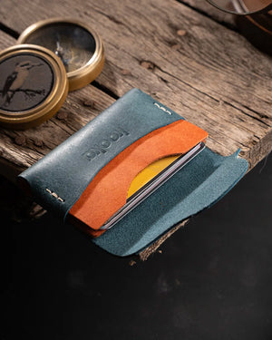 İki tonlu Full Grain (Üst Tabaka) Pueblo deri kartlık, slate mavi dış panel üzerinde deboss 'kooka' marka logosu ve kontrast turuncu/konyak iç kart cebi ile dikkat çekiyor; açık pozisyonda birden fazla kartın yelpaze gibi dışarı taştığı slim tasarım, el dikişi kenar işçiliği ve organik pull-up deri yüzeyindeki zengin renk derinliğiyle öne çıkıyor.