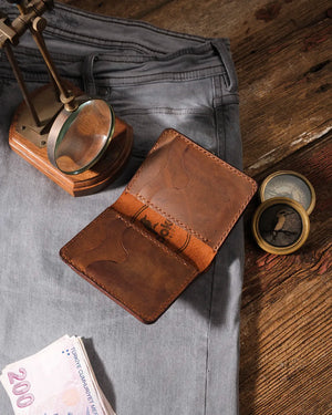 El dikişi deri cüzdan içi görünümü — OZN marka taba rengi hakiki deri bifold model, el dikişiyle açık pozisyonda; vintage pirinç pusula, büyüteç ve gri denim kumaş eşliğinde eskitilmiş ahşap zemin üzerinde düzenlenmiş.