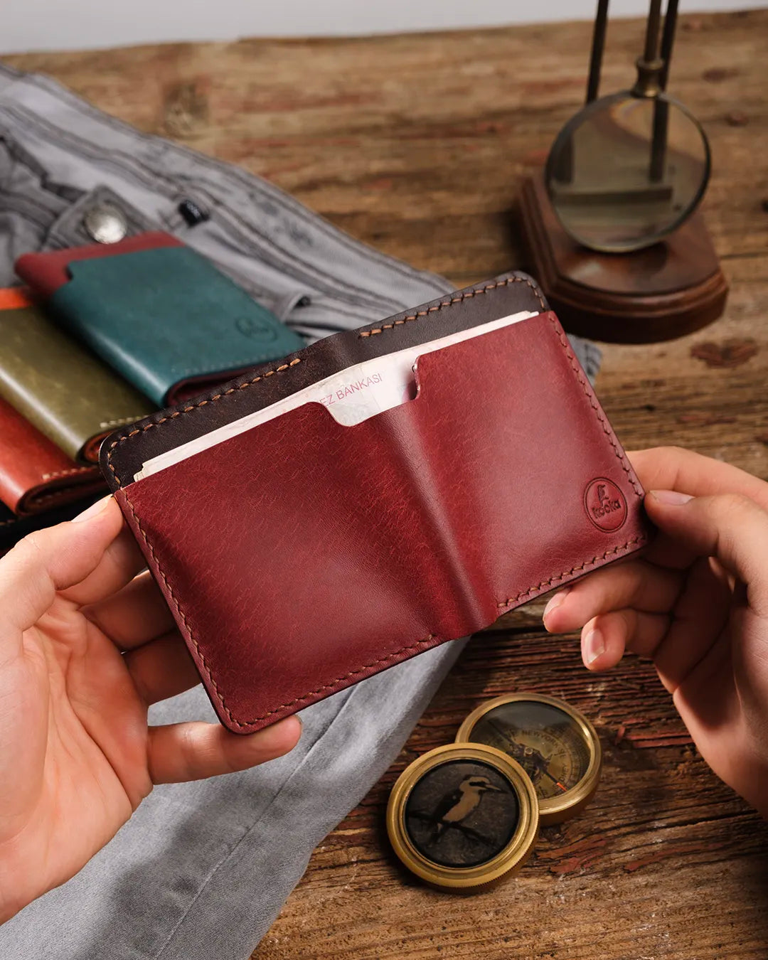 Elde tutulan açık bordo-kahverengi pueblo deri cüzdan, dış kısımda geniş para bölmesinde banknotlar görünüyor, Kooka Leather