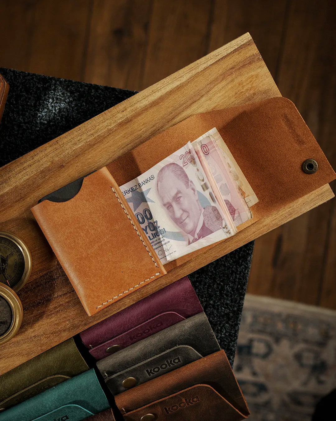 El dikişi deri cüzdan iç bölme detayı — camel rengi Pueblo derisiyle üretilen kooka STYLE bifold modeli açık konumda, Türk lirası banknotları nakit bölmesine yerleştirilmiş halde görünüyor; krem rengi el dikişi dikiş izleri yan panelde belirgin biçimde öne çıkıyor, pürüzsüz pull-up deri yüzeyi sıcak karamel tonlarıyla ışığı yansıtıyor. Arka planda serinin farklı renk seçenekleri — bordo, zeytin yeşili, koyu gri ve koyu kahverengi — sıralı şekilde yerleştirilmiş.