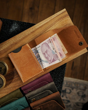 El dikişi deri cüzdan iç bölme detayı — camel rengi Pueblo derisiyle üretilen kooka STYLE bifold modeli açık konumda, Türk lirası banknotları nakit bölmesine yerleştirilmiş halde görünüyor; krem rengi el dikişi dikiş izleri yan panelde belirgin biçimde öne çıkıyor, pürüzsüz pull-up deri yüzeyi sıcak karamel tonlarıyla ışığı yansıtıyor. Arka planda serinin farklı renk seçenekleri — bordo, zeytin yeşili, koyu gri ve koyu kahverengi — sıralı şekilde yerleştirilmiş.