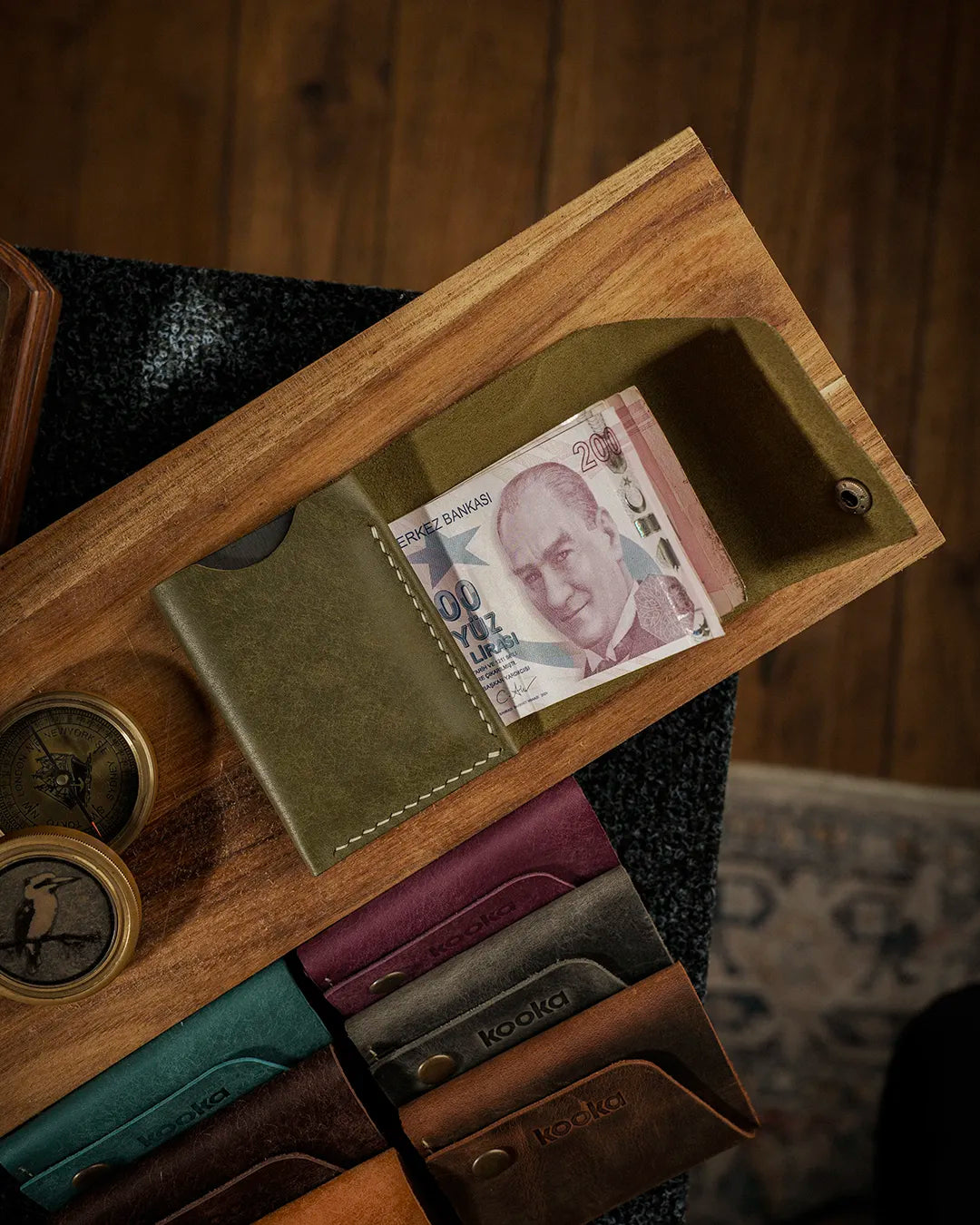 El dikişi deri cüzdan, zeytin yeşili Pueblo derisiyle üretilmiş kooka STYLE bifold modeli, açık konumda Türk lirası banknotlarını sergiliyor; krem renkli el dikişi dikiş detayı ve antik pirinç çıtçıt kapama görünür, matta balmumu yüzey dokusu ve doğal renk geçişleriyle öne çıkan premium işçilik göze çarpar; arka planda serinin tüm renkleri sıralanmış