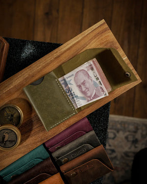 El dikişi deri cüzdan, zeytin yeşili Pueblo derisiyle üretilmiş kooka STYLE bifold modeli, açık konumda Türk lirası banknotlarını sergiliyor; krem renkli el dikişi dikiş detayı ve antik pirinç çıtçıt kapama görünür, matta balmumu yüzey dokusu ve doğal renk geçişleriyle öne çıkan premium işçilik göze çarpar; arka planda serinin tüm renkleri sıralanmış