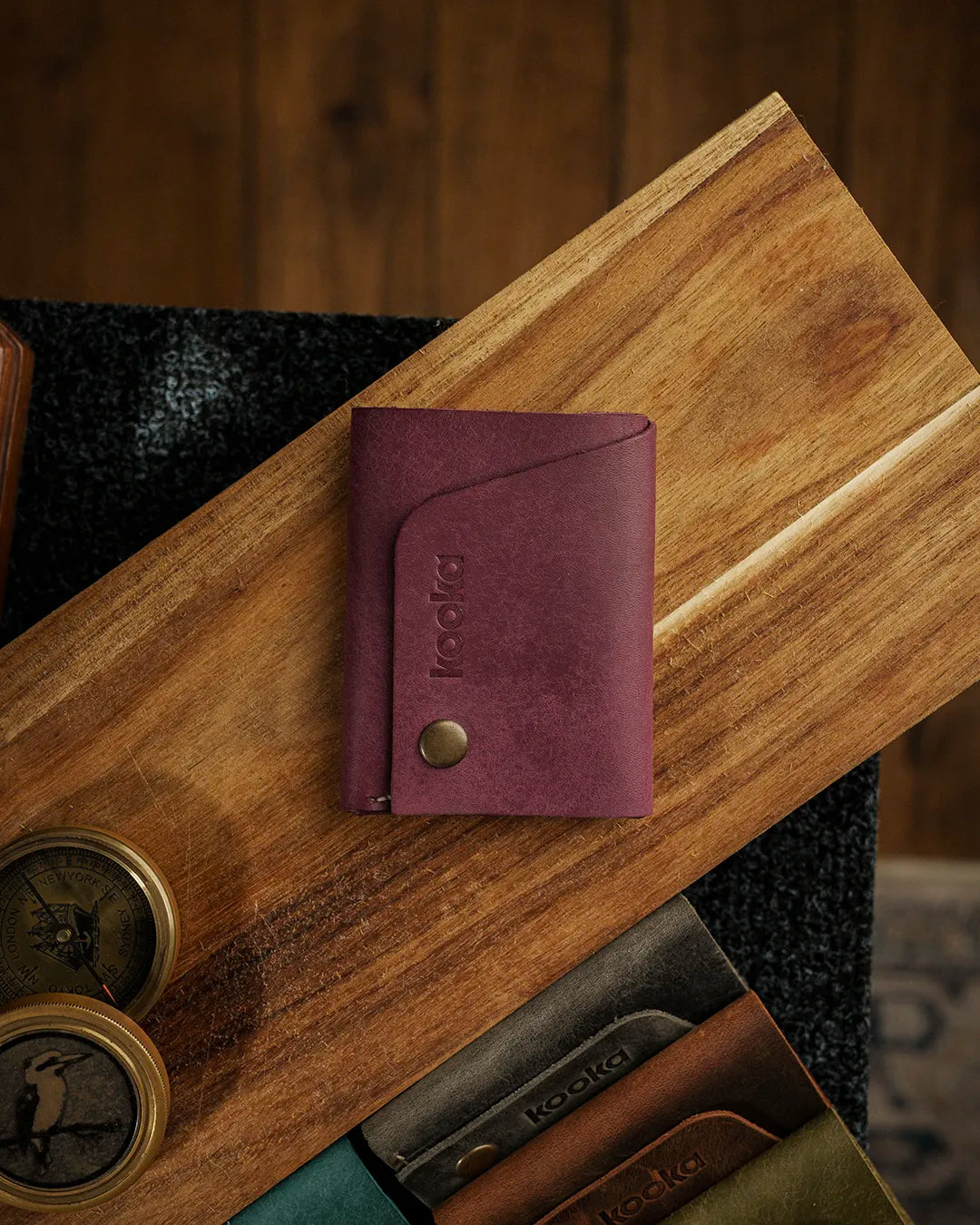 El dikişi deri cüzdan kapaklı kapatma detayı — kooka STYLE kompakt bifold cüzdan, Pueblo deri mor/mürdüm rengi, mat ve hafif balmumu dokusunda yüzey, antik pirinç çıtçıt düğme kapama, kabartma kooka logo ve temiz yanmış kenar işçiliğiyle üst açıdan düz yatay çekim.