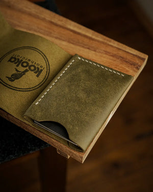 El dikişi deri cüzdan, zeytin yeşili Pueblo derisiyle üretilmiş kooka STYLE bifold model; matta, mumlu yüzey dokusu ve krem rengi el dikişi dikişleriyle ön yüzey detayı, kart bölmesinden hafifçe görünen kartlarla kapalı konumda üstten çekim.