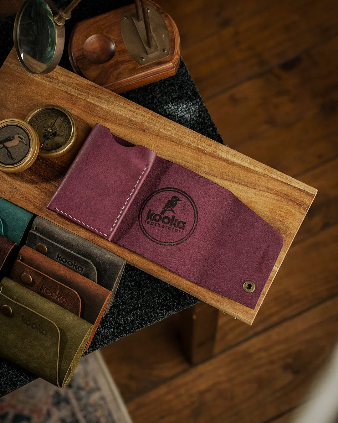 El dikişi mor Pueblo deri bifold cüzdan, açık kapaklı düz fotoğraf; cüzdanın iç yüzündeki süet dokulu mor deri yüzey, emboss baskılı kooka logotype ve antik pirinç çıtçıt kapama detayıyla birlikte, yanda sıralanan zeytin yeşili, koyu kahverengi ve teal renk seçenekleriyle tam koleksiyon görünümü