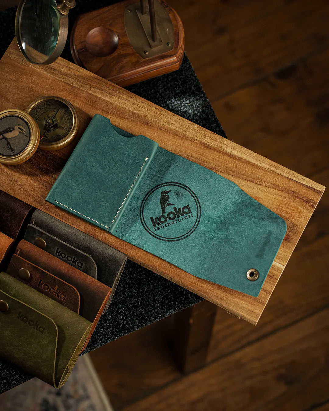 Vintage karakterli Pueblo deri bifold cüzdan, teal rengi mavi-yeşil tonlarında pull-up yüzey dokusuyla kapalı pozisyonda üstten görünüm; ön yüzeyde belirgin kooka Leathercraft marka logosu, antik pirinç çıtçıt kapama ve soldan dikkat çeken krem renkli el dikişi; yanında aynı seriden kahverengi, koyu gri, zeytin yeşili ve taba rengi cüzdanlarla tam koleksiyon sunumu