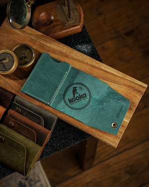 Vintage karakterli Pueblo deri bifold cüzdan, teal rengi mavi-yeşil tonlarında pull-up yüzey dokusuyla kapalı pozisyonda üstten görünüm; ön yüzeyde belirgin kooka Leathercraft marka logosu, antik pirinç çıtçıt kapama ve soldan dikkat çeken krem renkli el dikişi; yanında aynı seriden kahverengi, koyu gri, zeytin yeşili ve taba rengi cüzdanlarla tam koleksiyon sunumu