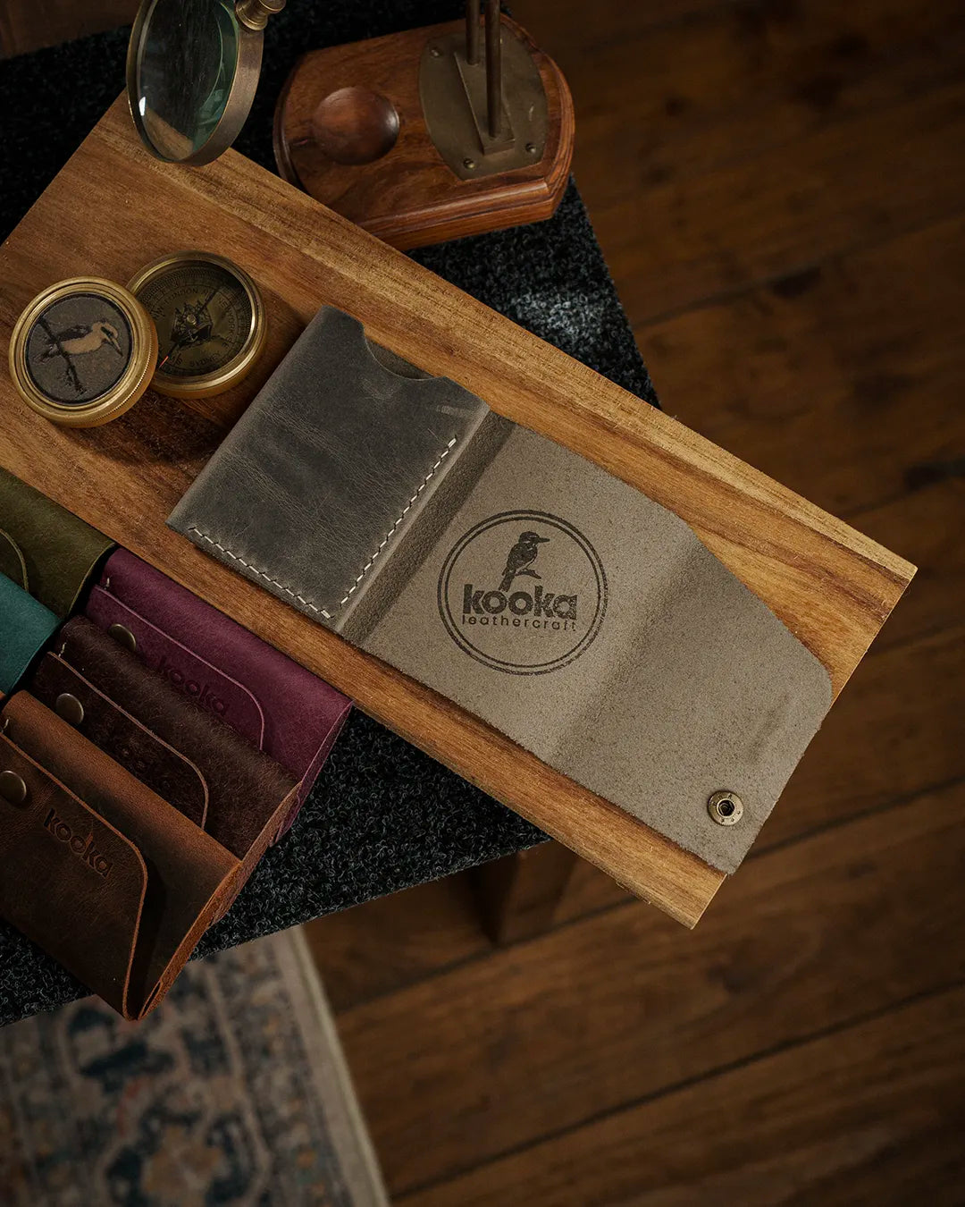 Full Grain (Üst Tabaka) Crazy Deri bifold cüzdanın gri/zeytin tonundaki versiyonu öne çıkarılmış, kooka leathercraft marka logosu lazer kazıma ile işlenmiş, el dikişi detayı ve antik pirinç çıtçıt kapama görünür halde; serinin tüm renk seçenekleri olan koyu kahve, bordo, yeşil ve turkuaz tonları arka planda sıralanmış, distressed mat yüzey dokusu ve doğal çiziklerle zenginleştirilmiş el yapımı işçilik kalitesi ön plana taşınmış.