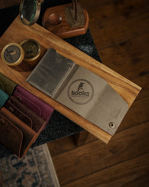 Full Grain (Üst Tabaka) Crazy Deri bifold cüzdanın gri/zeytin tonundaki versiyonu öne çıkarılmış, kooka leathercraft marka logosu lazer kazıma ile işlenmiş, el dikişi detayı ve antik pirinç çıtçıt kapama görünür halde; serinin tüm renk seçenekleri olan koyu kahve, bordo, yeşil ve turkuaz tonları arka planda sıralanmış, distressed mat yüzey dokusu ve doğal çiziklerle zenginleştirilmiş el yapımı işçilik kalitesi ön plana taşınmış.