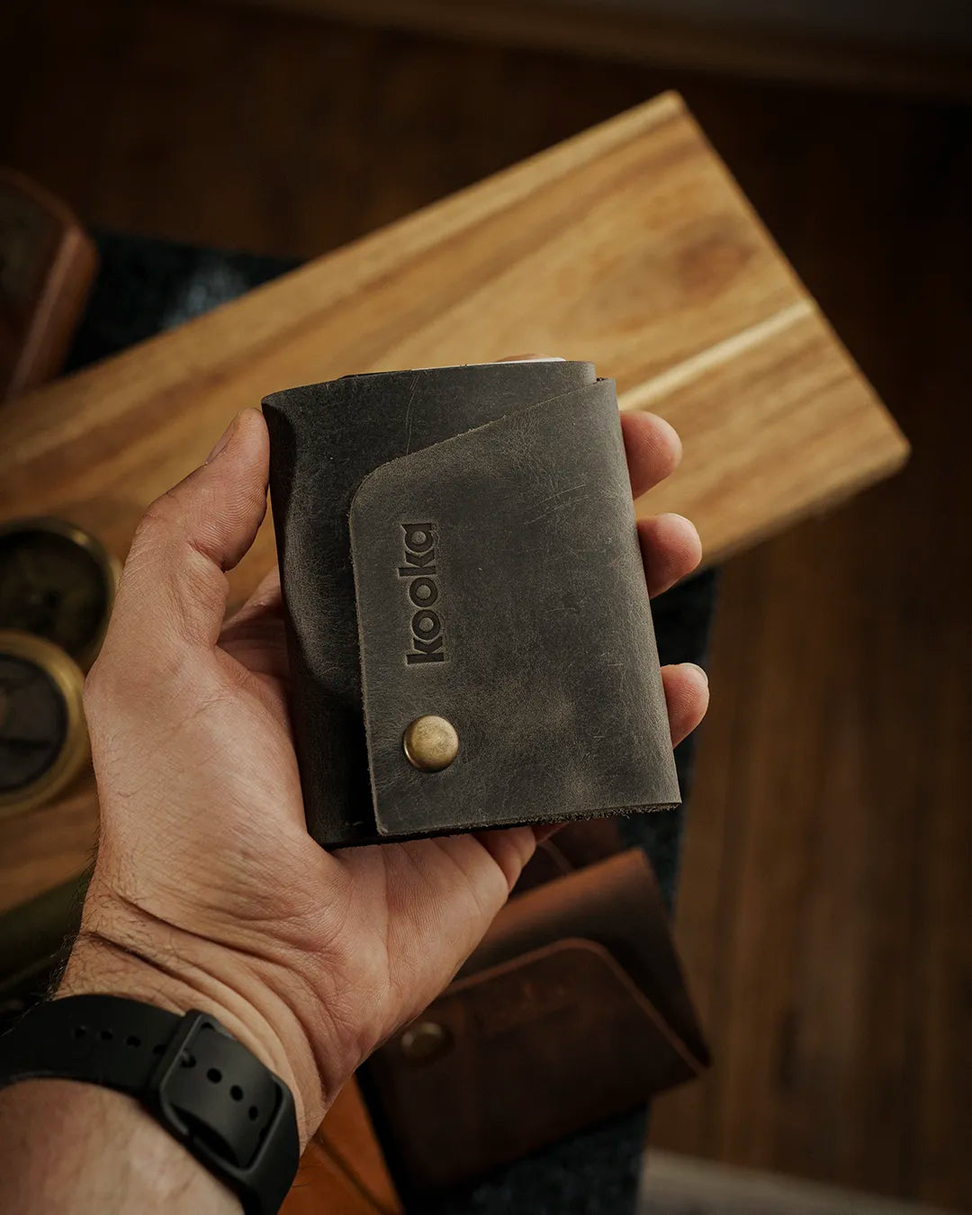 Crazy Deri kompakt bifold cüzdan, erkek avucunda sergileniyor; koyu gri-haki rengi, distressed mat yüzeyi ve doğal çizgi izleriyle zengin doku sunan Full Grain (Üst Tabaka) deri kullanımı öne çıkıyor. Kooka Style dikey deri cüzdanın ön yüzünde lazer kabartmalı 'kooka' logosu ve antik pirinç çıtçıt kapama düğmesi belirgin şekilde görülüyor; köşegen kart bölmesi kapağı tasarımı ve sağlam el işçiliği dikkat çekiyor.